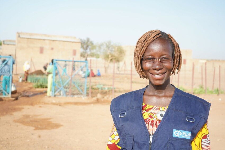 Agricultural coordinator Nikiema, wearing her Plan International jacket,  smiles in front of the school garden, 