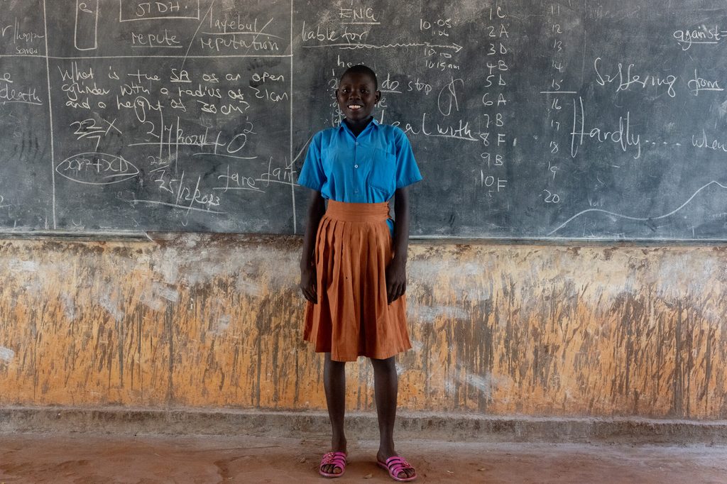 Winnie, 14, stands by the blackboard in her school with a smile on her face.