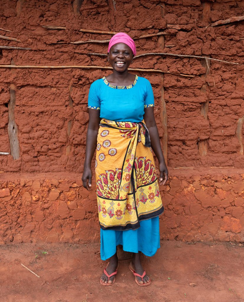 Khadijah stands against the brick red of the school. She is wearing brightly coloured clothes and is smiling. 