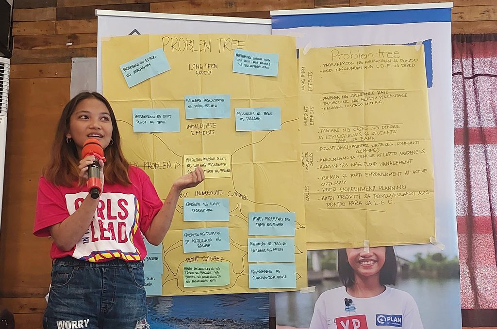 Aires standing in front of a poster board where her problem tree is drawn speaking into a microphone. 