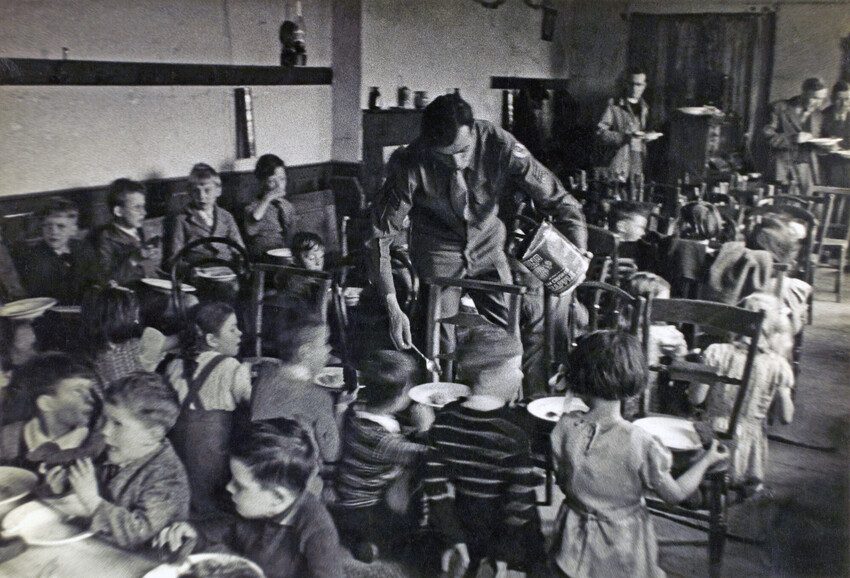 An officer serves food to the children sitting at the table. Black and white photo 1944. 