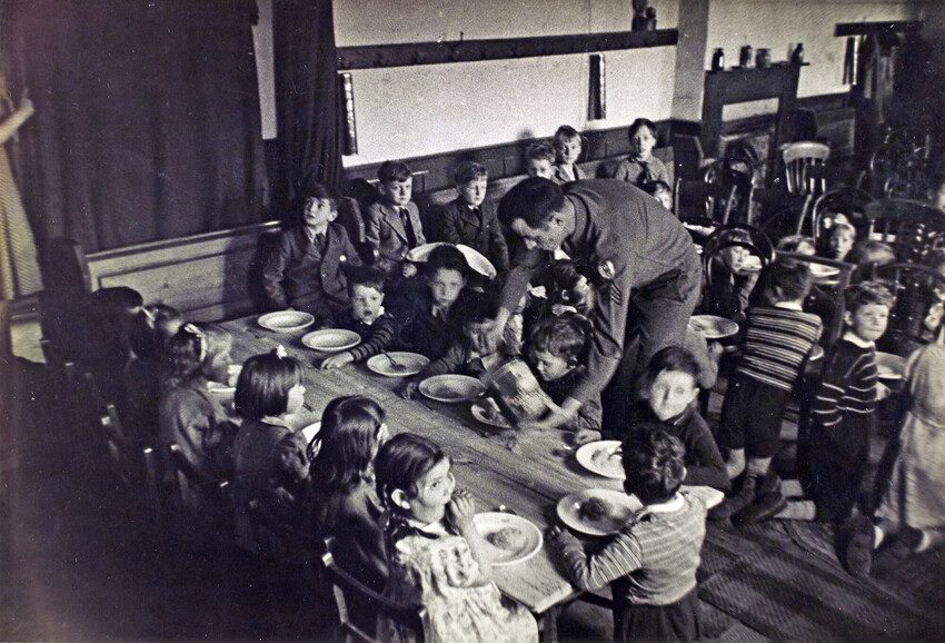 An officer serves food to the children sitting at the table. Black and white photo 1944. 