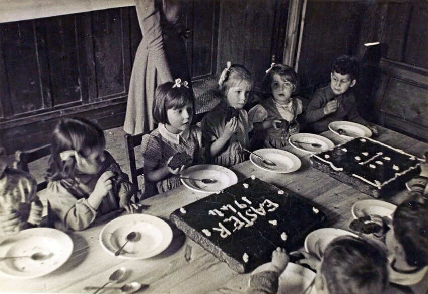 A black and white photo from 1944 of girls and boys sitting around a bench table eating cake. 