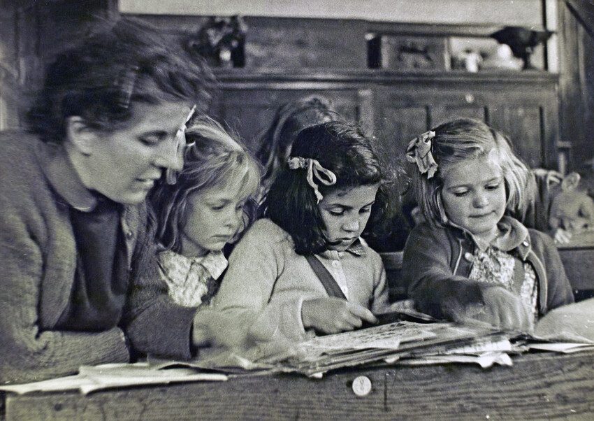 Black and white photo 1944. 3 girls do school work with their teacher at a desk.