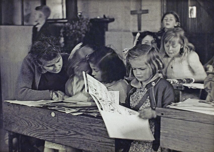 Black and white photo 1944. A young girl reads at a desk next to 2 other girls who are talking with their teacher. 