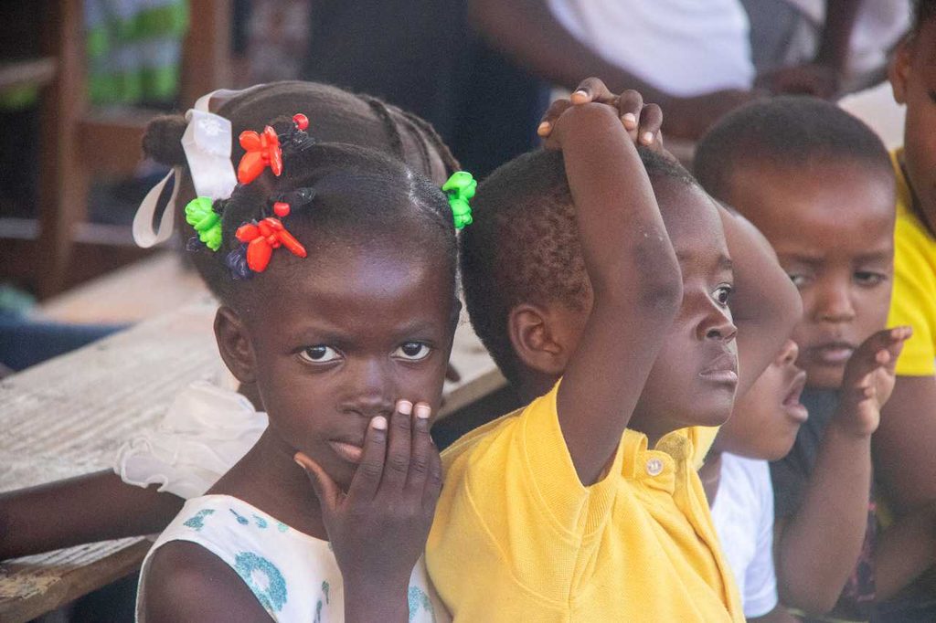 A young girl faces the camera, she is sitting on a bench with other children.