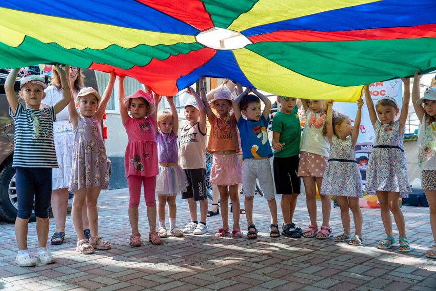 Lots of children lift up a multi-coloured play parachute. 