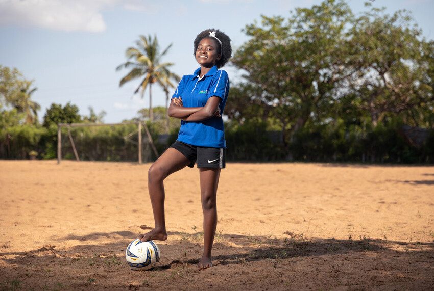 Eunice stands with her foot on her football, with her arms folded, smiling. 