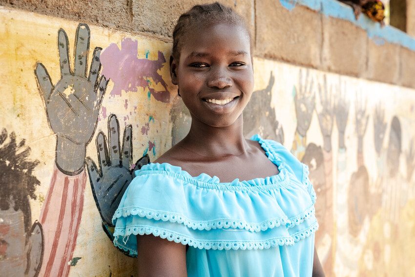 Nyabhan, a 19 year old girl from South Sudan, now living in Ethiopia stands against her school wall and smiles at the camera.