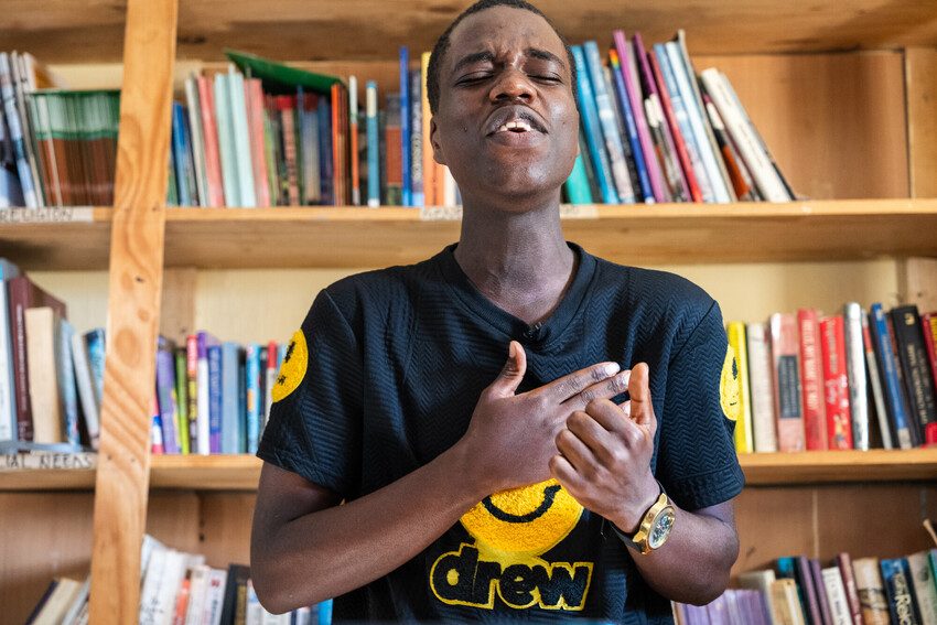 Heritier, a young refugee activist performs a poem in front of a bookcase.