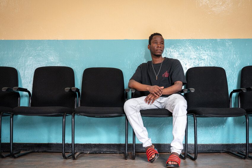 Damien, a refugee youth activist and humanitarian sits with hands folded, looking pensively to his left.