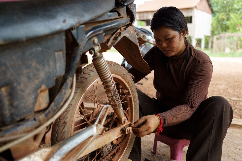 Kanada kneels down repairing the back of a motorbike. 