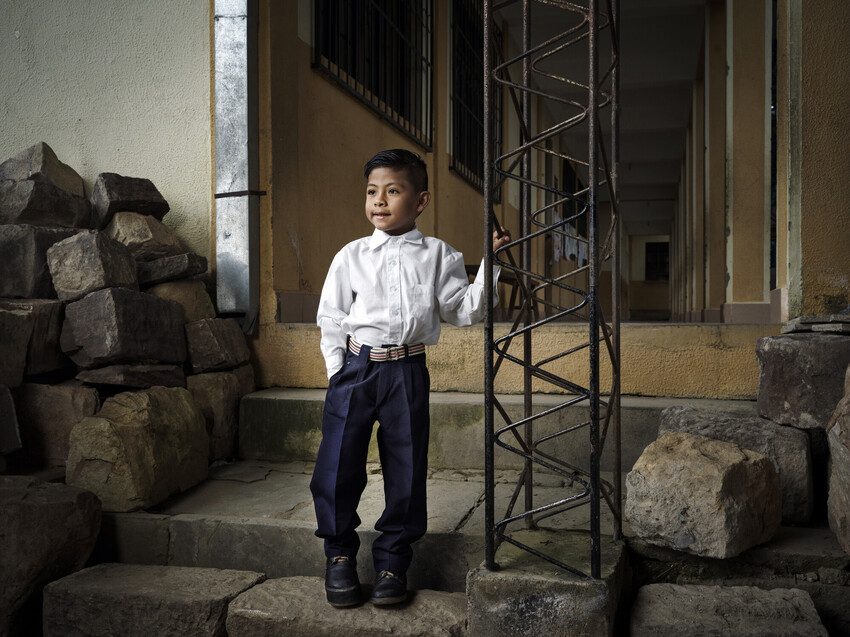 Six-year-old Gael  stands on the steps to his classroom. It is rocky and uneven so he holds on to some railings for support.
