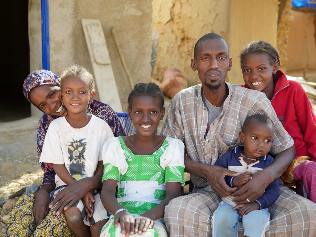 Aissata sits in front of her house with her husband and their children.