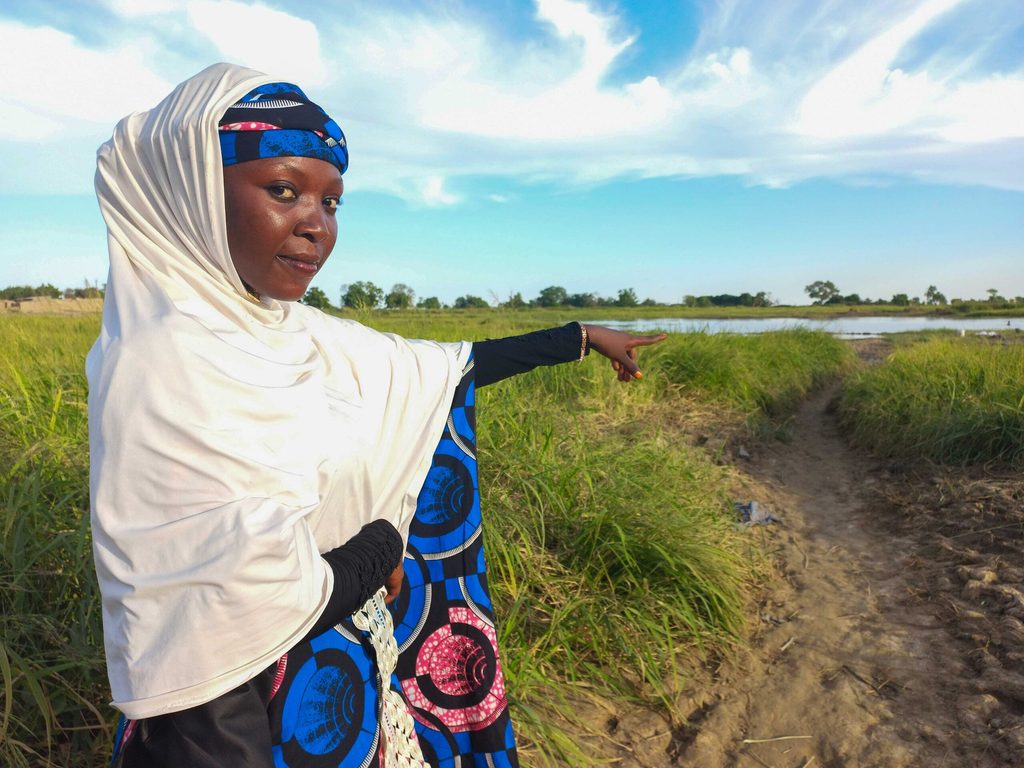 Halima stands in a grassy area near the Yobe River in north-eastern Nigeria, pointing toward a flood-prone pathway as she explains changes in the landscape following seasonal flooding.