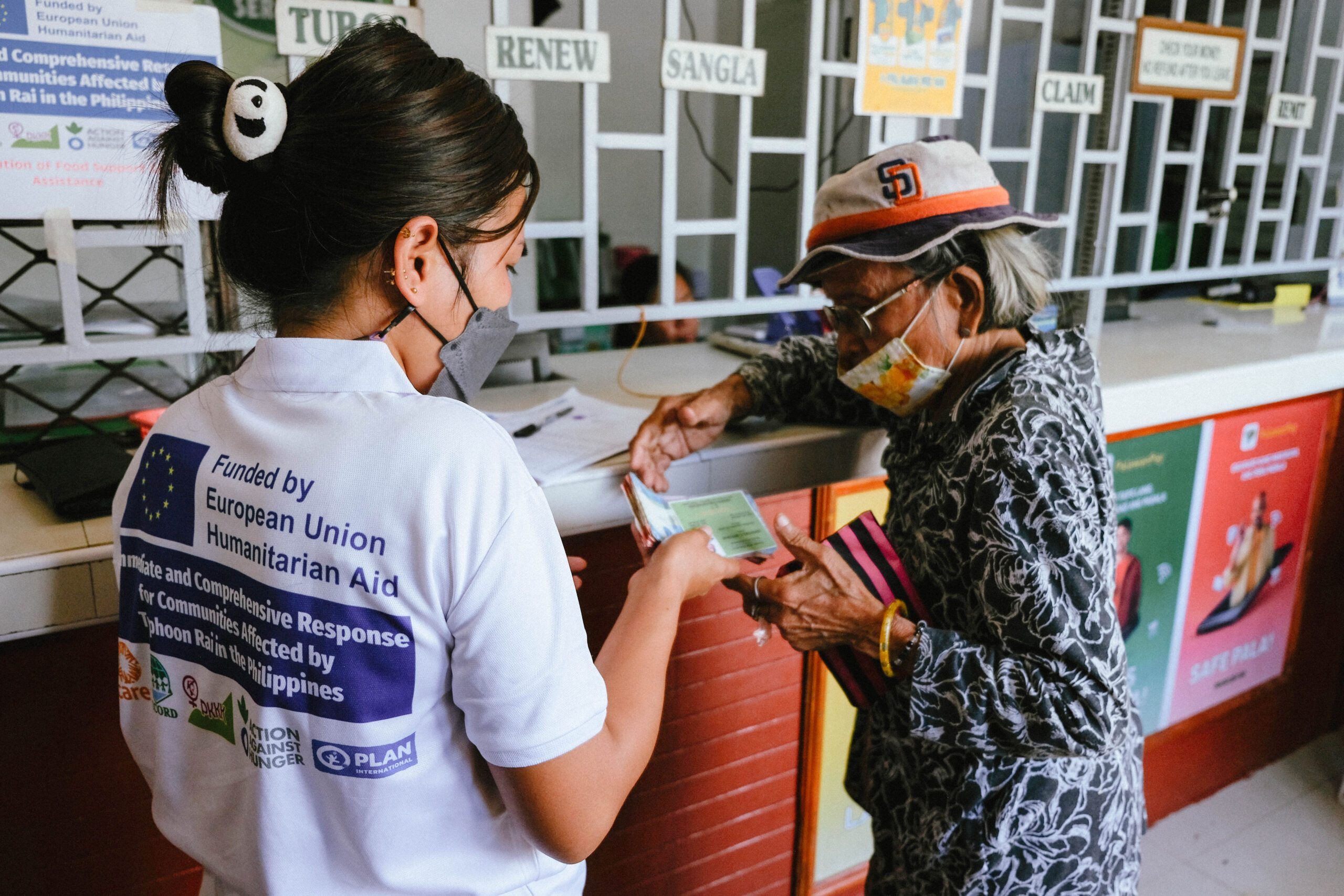A Plan International staff member assists an elderly woman in obtaining cash for food assistance