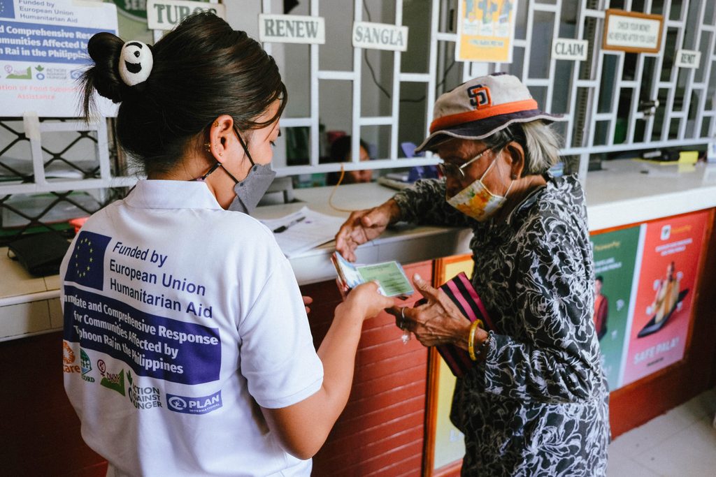 A Plan International staff member assists an elderly woman in obtaining cash for food assistance 