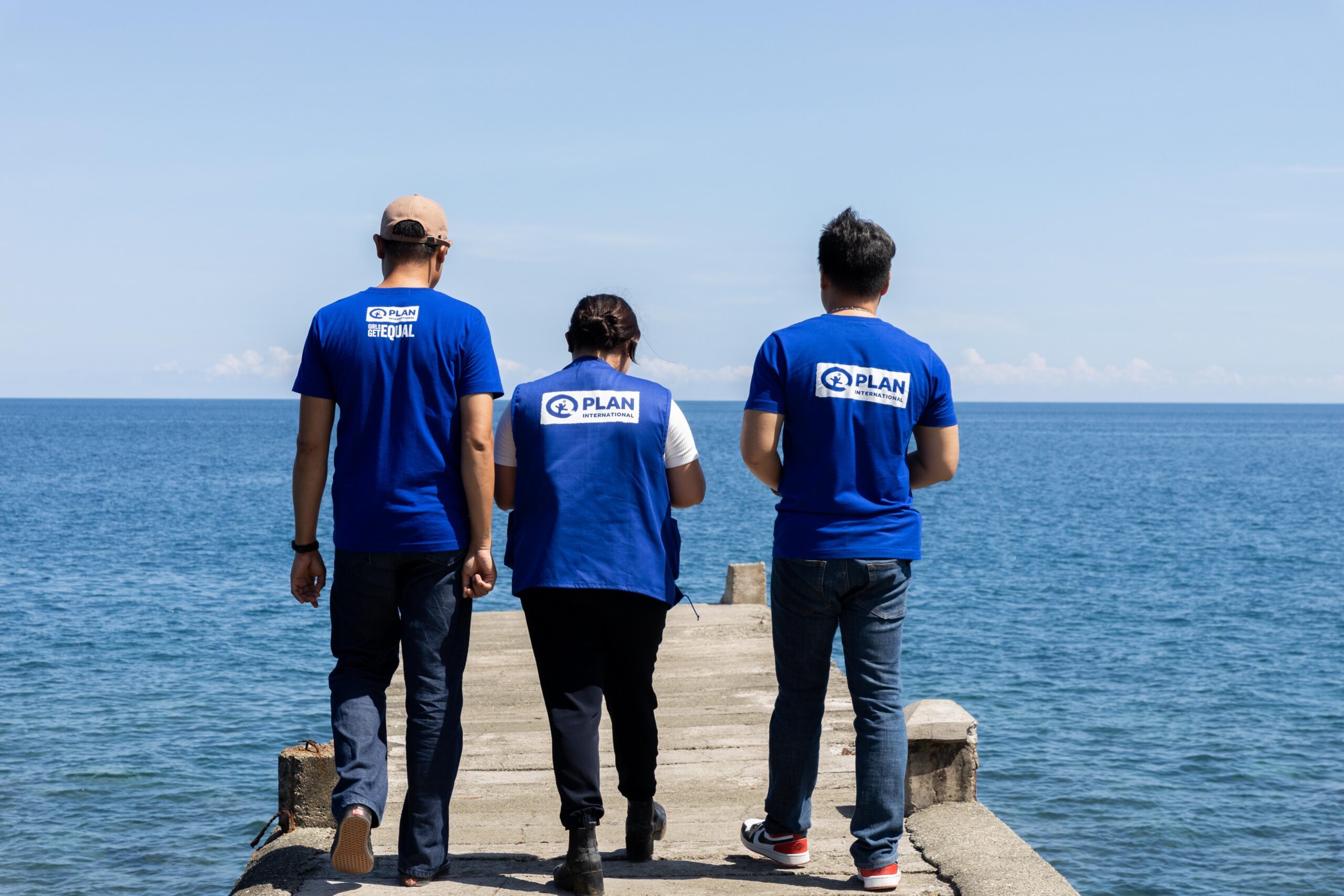 Three Plan International staff members stand on a board walk above the sea in Plan International branded clothing