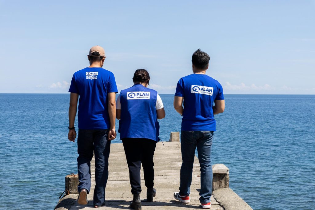 Three Plan International staff members stand on a board walk above the sea in Plan International branded clothing