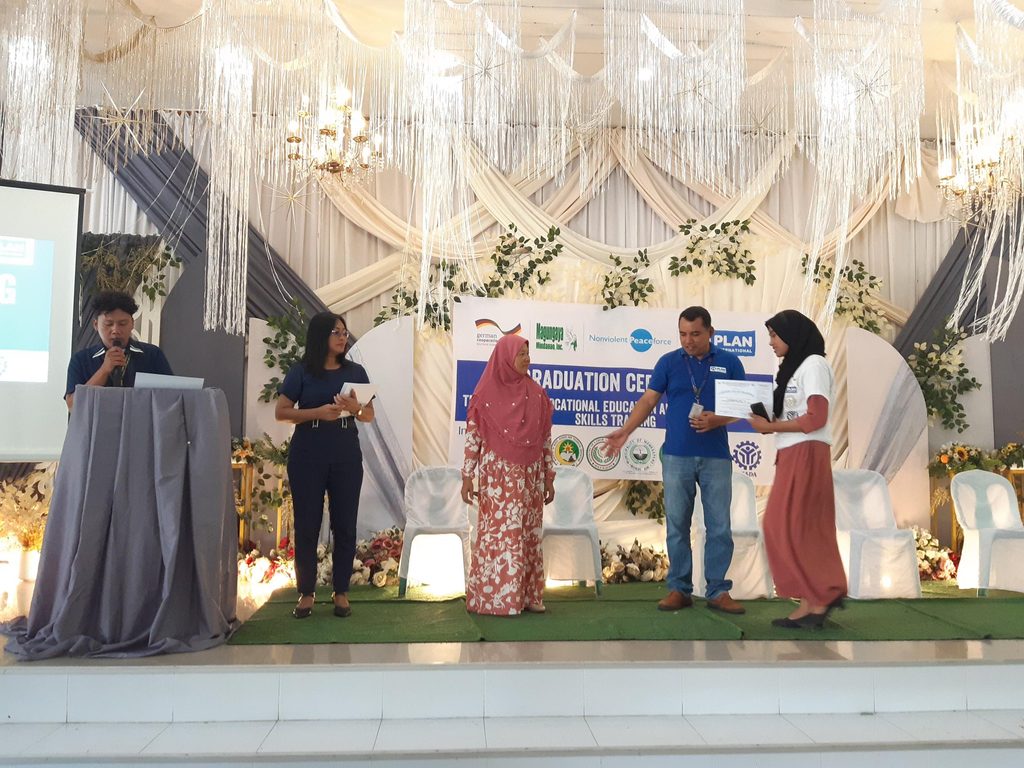 Four adults stand on stage as a young female graduate receives her diploma