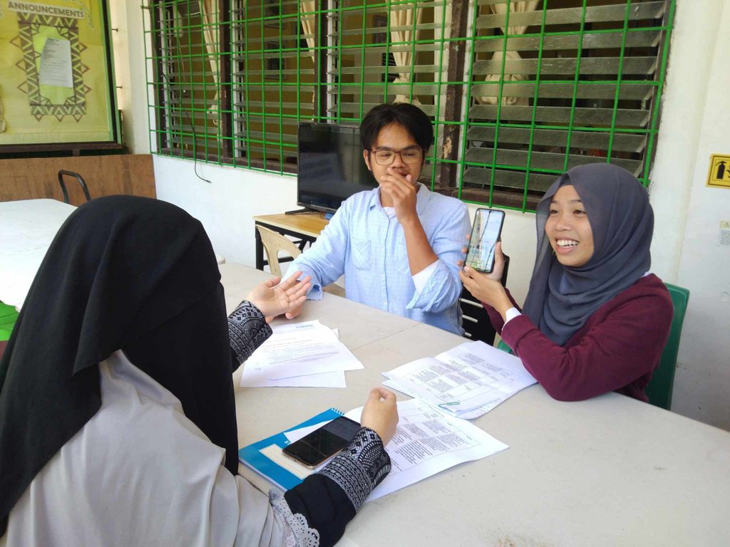 A group of 3 young people are seated around a table, discussing a smartphone app