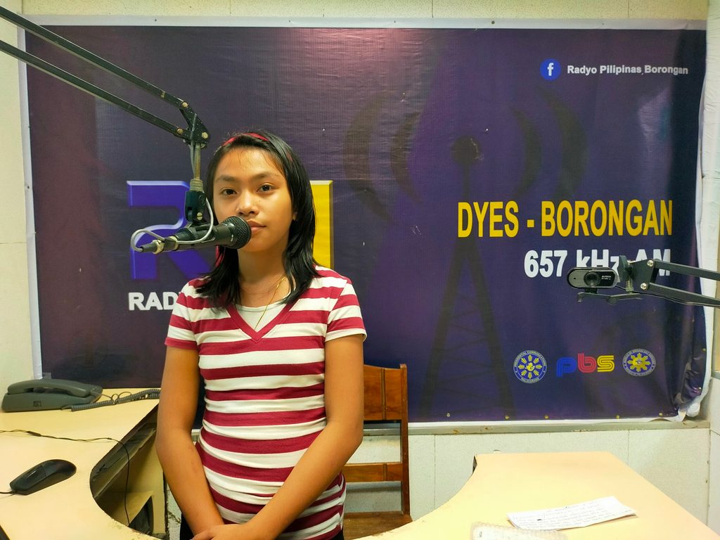A young girl sits in a radio booth behind a microphone