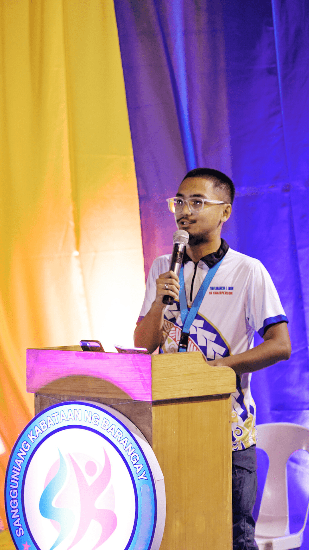 The person in the photo is standing behind a wooden podium with a lit emblem of the Sangguniang Kabataan on the front. They are holding a microphone in one hand, speaking or addressing an audience. They are wearing clear eyeglasses, a white sports-style shirt with blue and yellow designs, and a lanyard around their neck. The background features bright yellow, purple, and blue drapes, creating a vibrant stage setting.