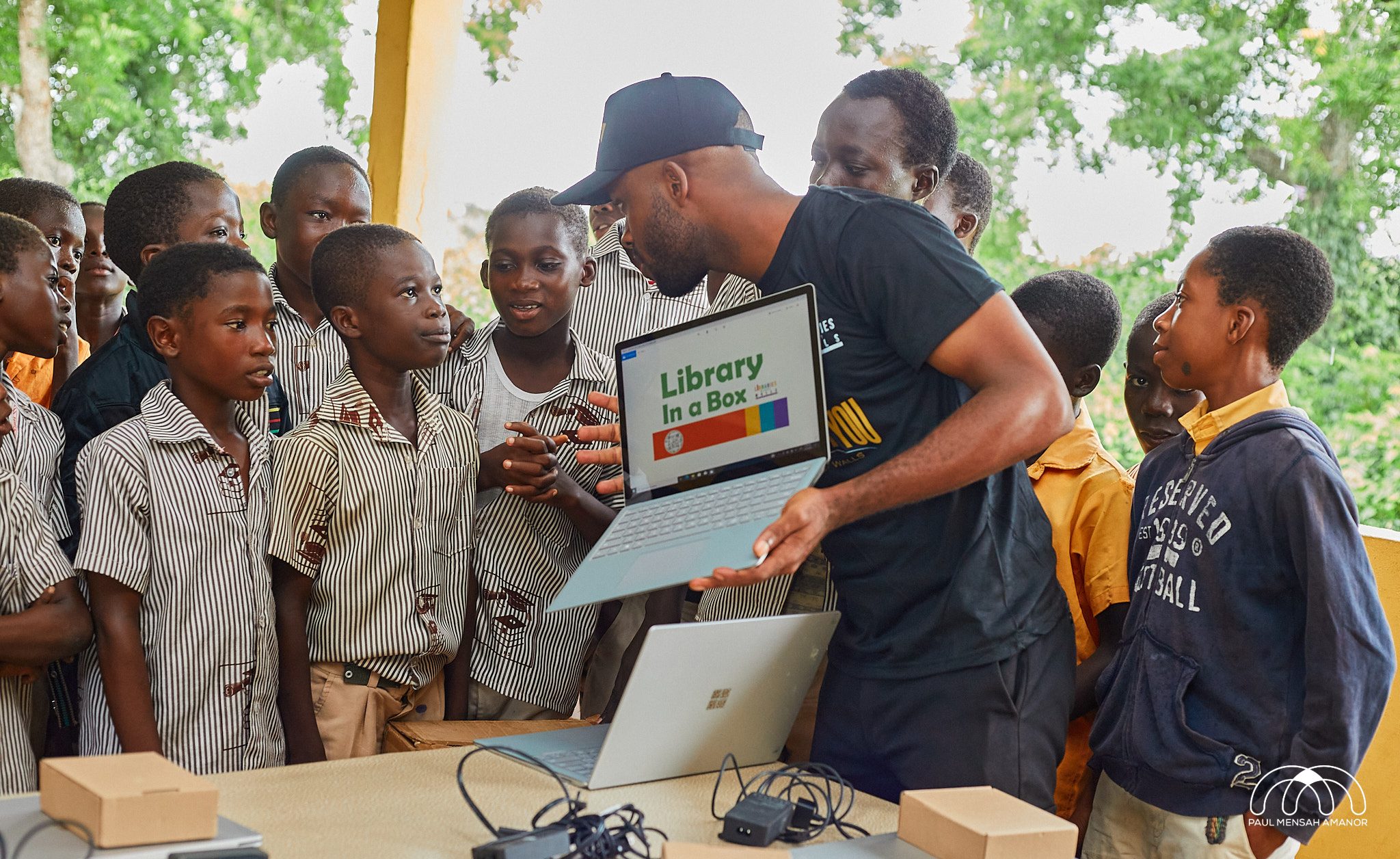 A group of young children gathered around a laptop with the words "Library in a box"