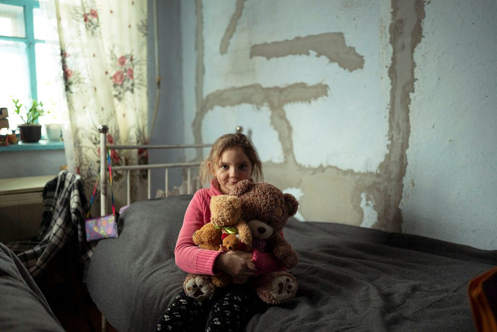 Young girl in Ukraine sitting on her bed, holding her teddy bears.