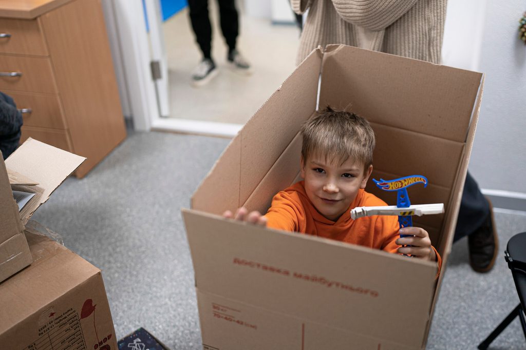 A young boy in Ukraine playing inside a box while at school, showing his toy at the camera.