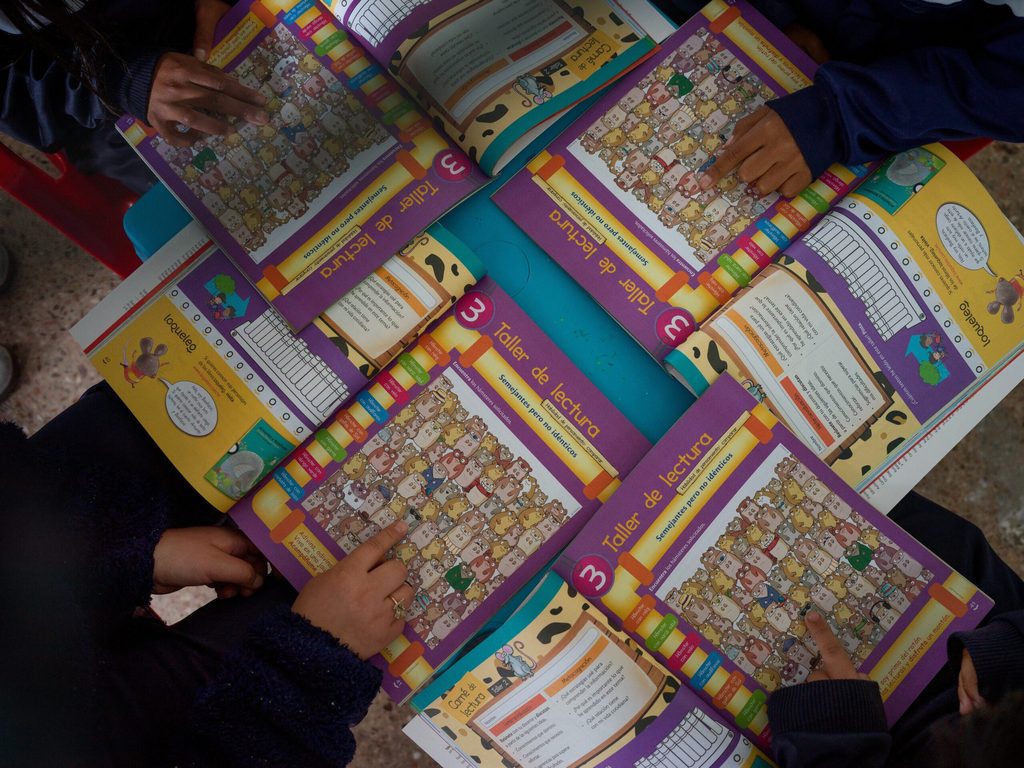 Several text books laying on a desk in a classroom while children are reading through them.