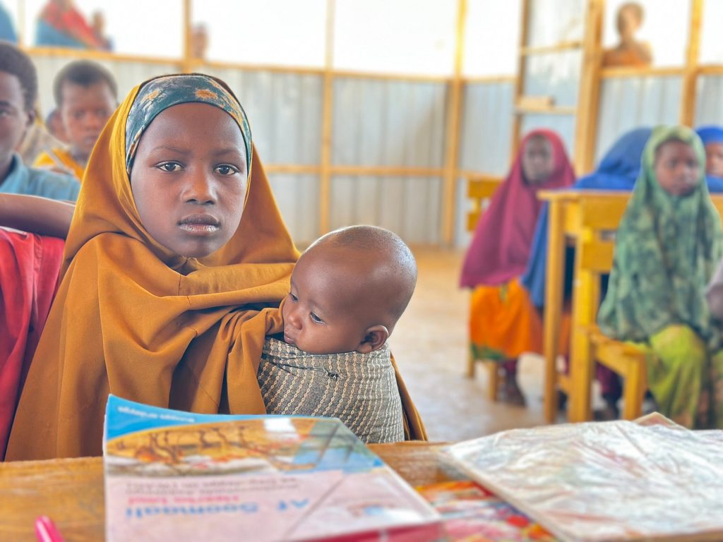 A girl sitting by a classroom desk while holding her 6-month-old sister. A school book is in front of her on the desk and classmates are seen around her sitting on their chairs.