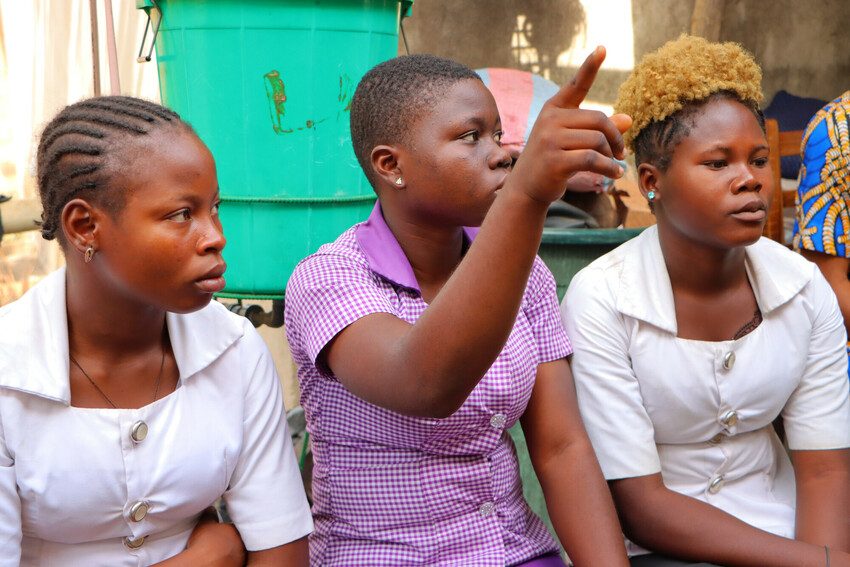 Majoie sitting between two other young women holding up her hand to ask a question. 