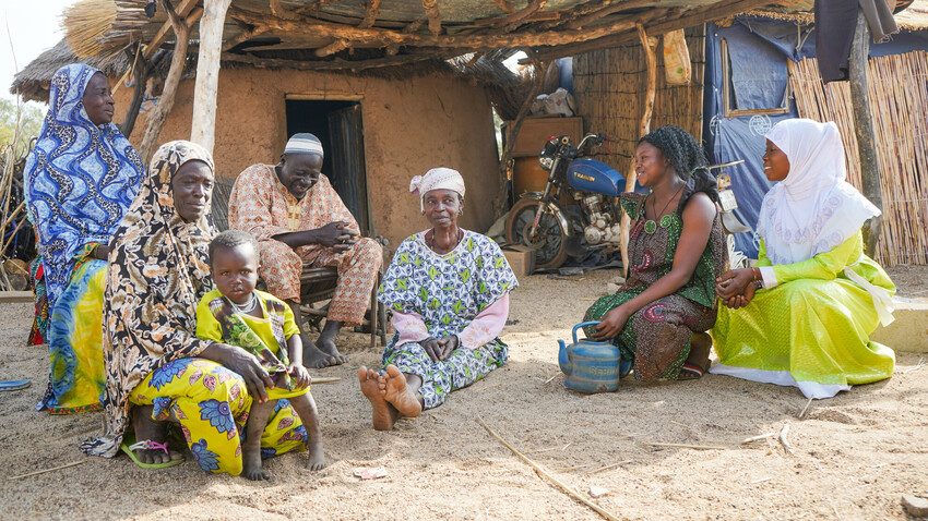 Bernadette visiting her friend's family living in a displacement camp. They are sitting in front of their home. 