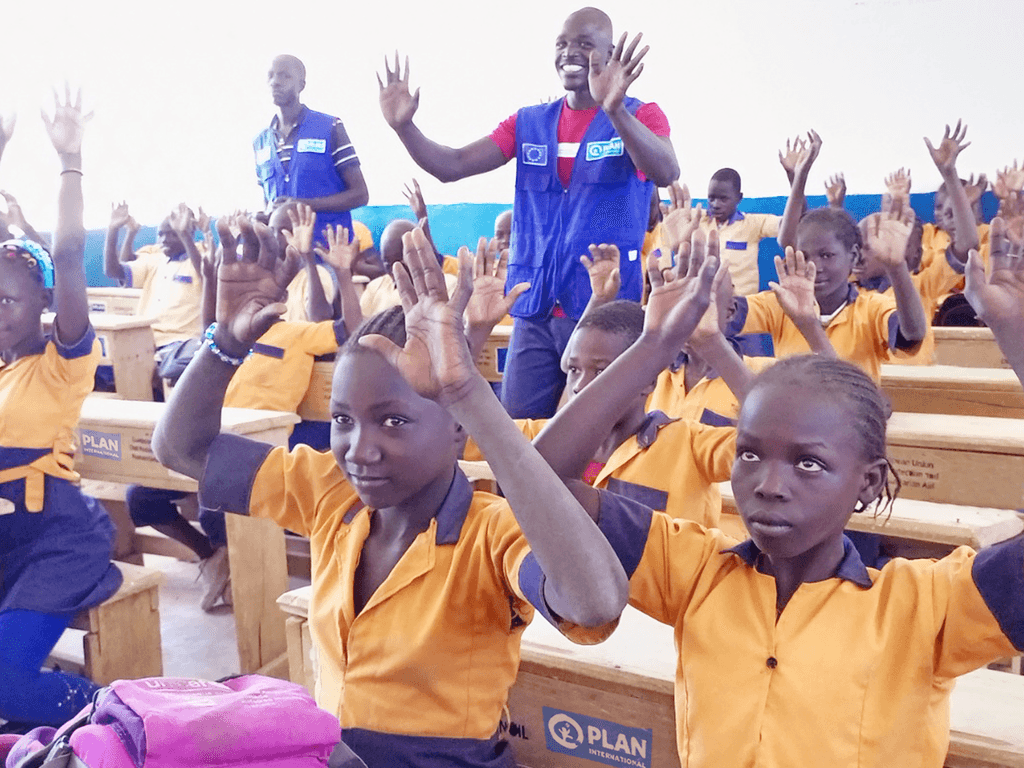 Children in a classroom hold their arms up in the air.