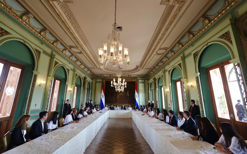 A long table with all the girl ministers sitting and looking towards Adriana who sits at the top of the table with the President.