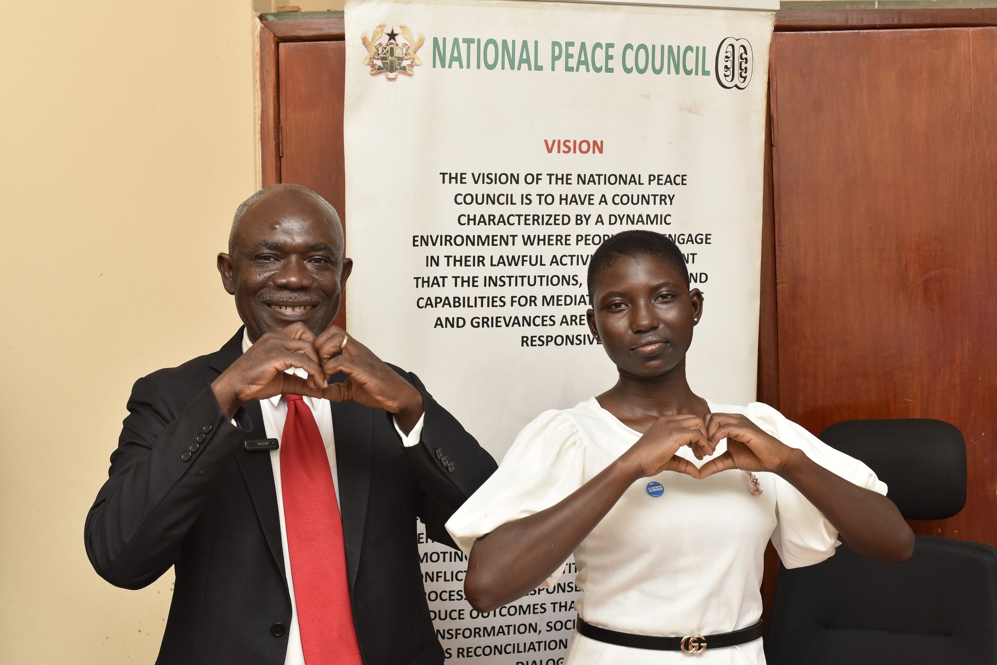 Sophia and the Board Chair of the National Peace Council make the #uniteforpeace heart symbols with their hands. 