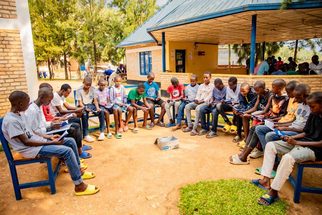 Children sit in a circle on chairs outside during an education session.