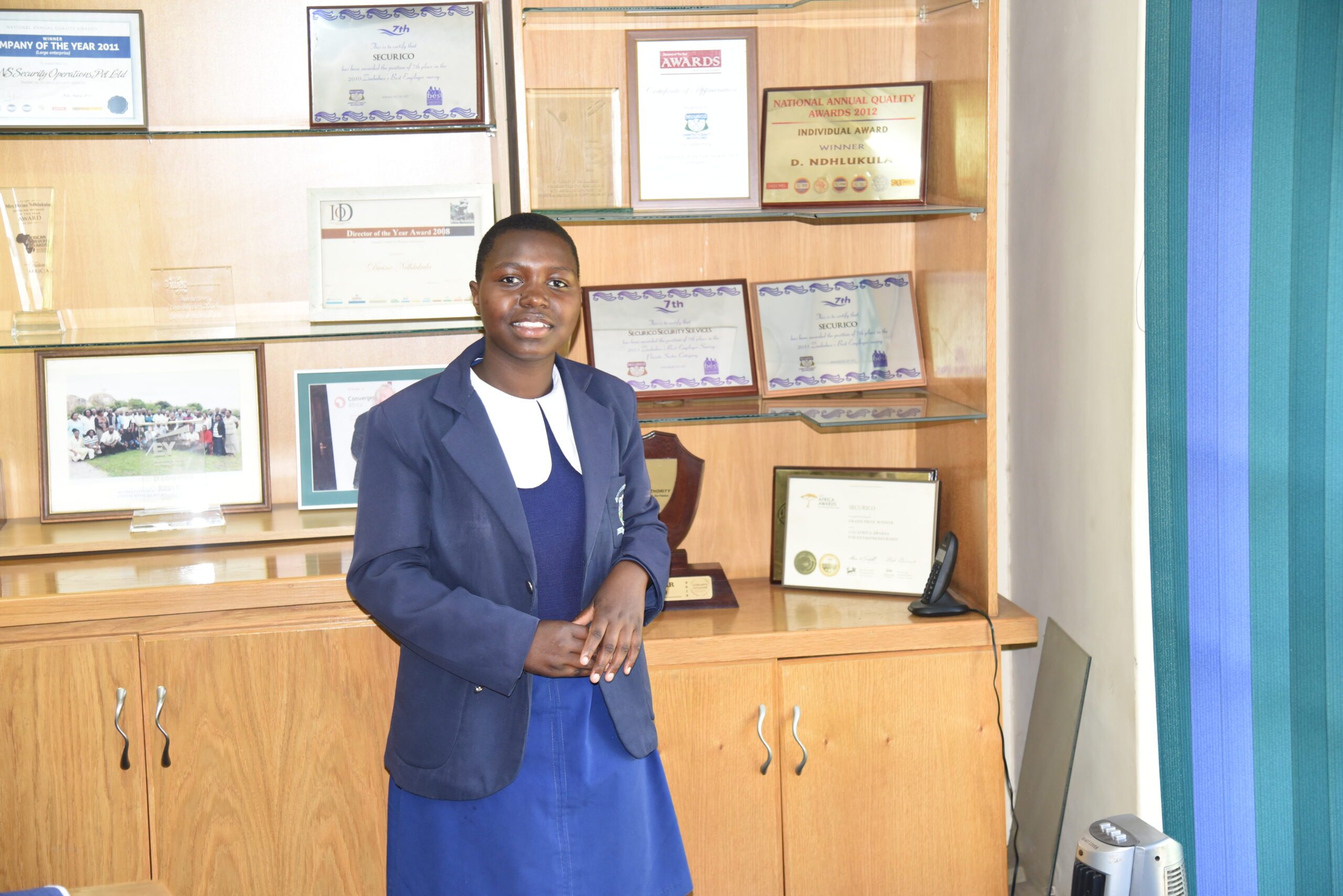 Delisa (15) posing for a picture in front of numerous accolades of excellency won by Securico Managing Director Divine Simbi Ndhlukula