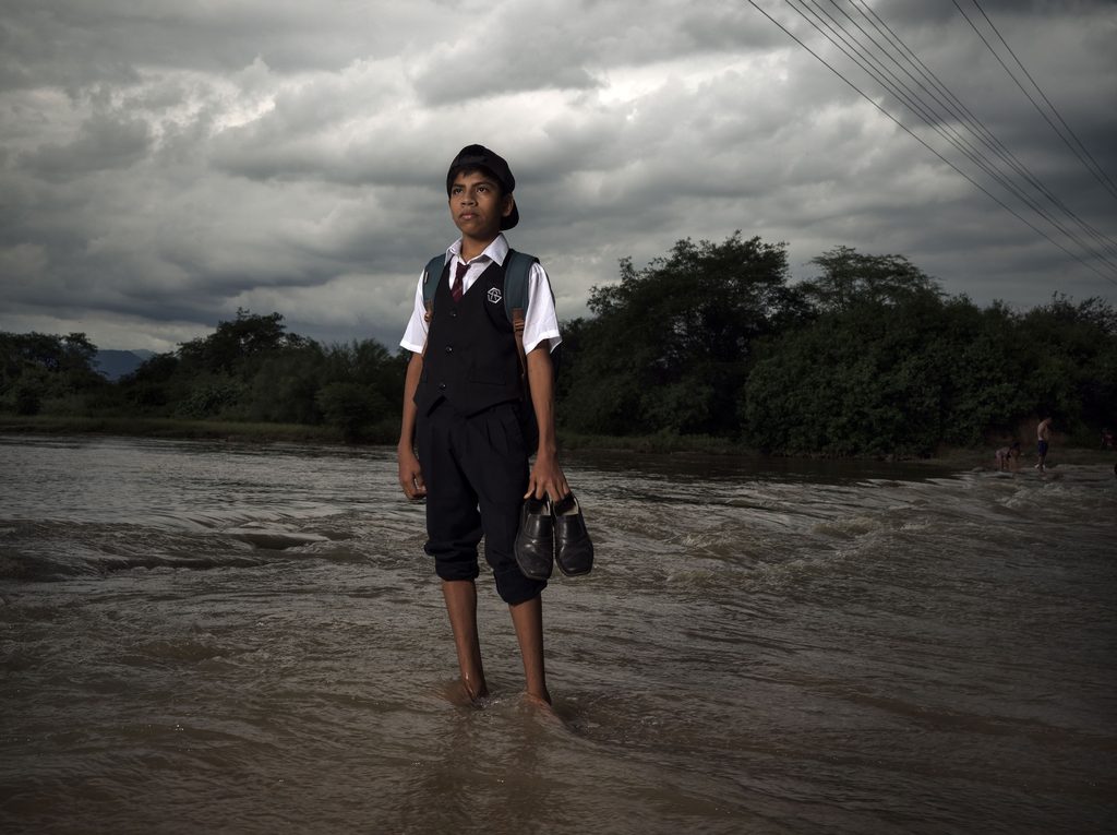 Stalin (14) from Piura, Peru stands at the edge of the overflowing river in his school uniform, determined yet uncertain—each flood season turns the simple act of going to school into a daily risk. © Plan International / Chris de Bode.