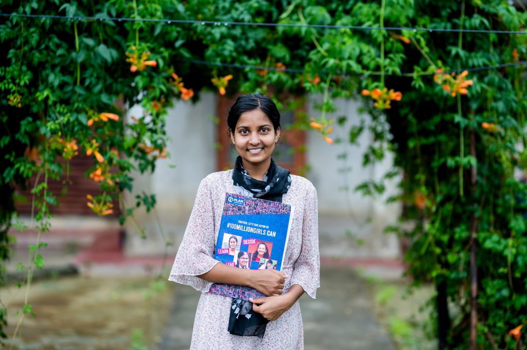 Rahimat standing in front of a gate, smiling and holding a book.