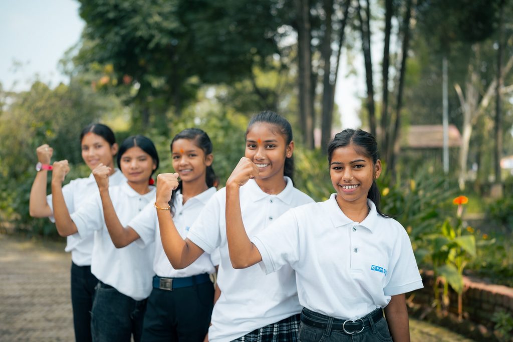 Five girls are raising their arms and smiling at the camera.