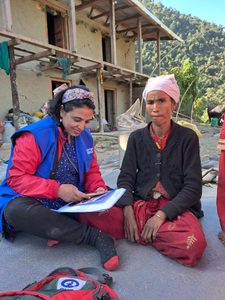 Kabita is wearing blue jacket with Plan's logo sitting on the floor and talking to women wearing black sweater and red sari.