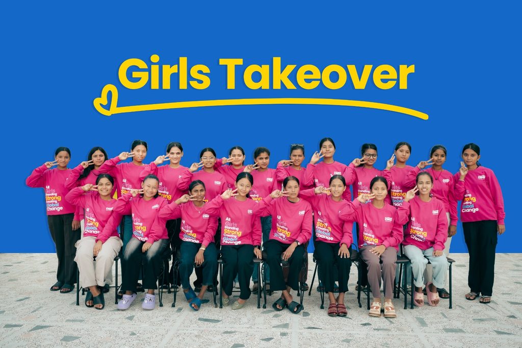 25 girls wearing pink sweatshirt and posing with equal sign with their fingers.