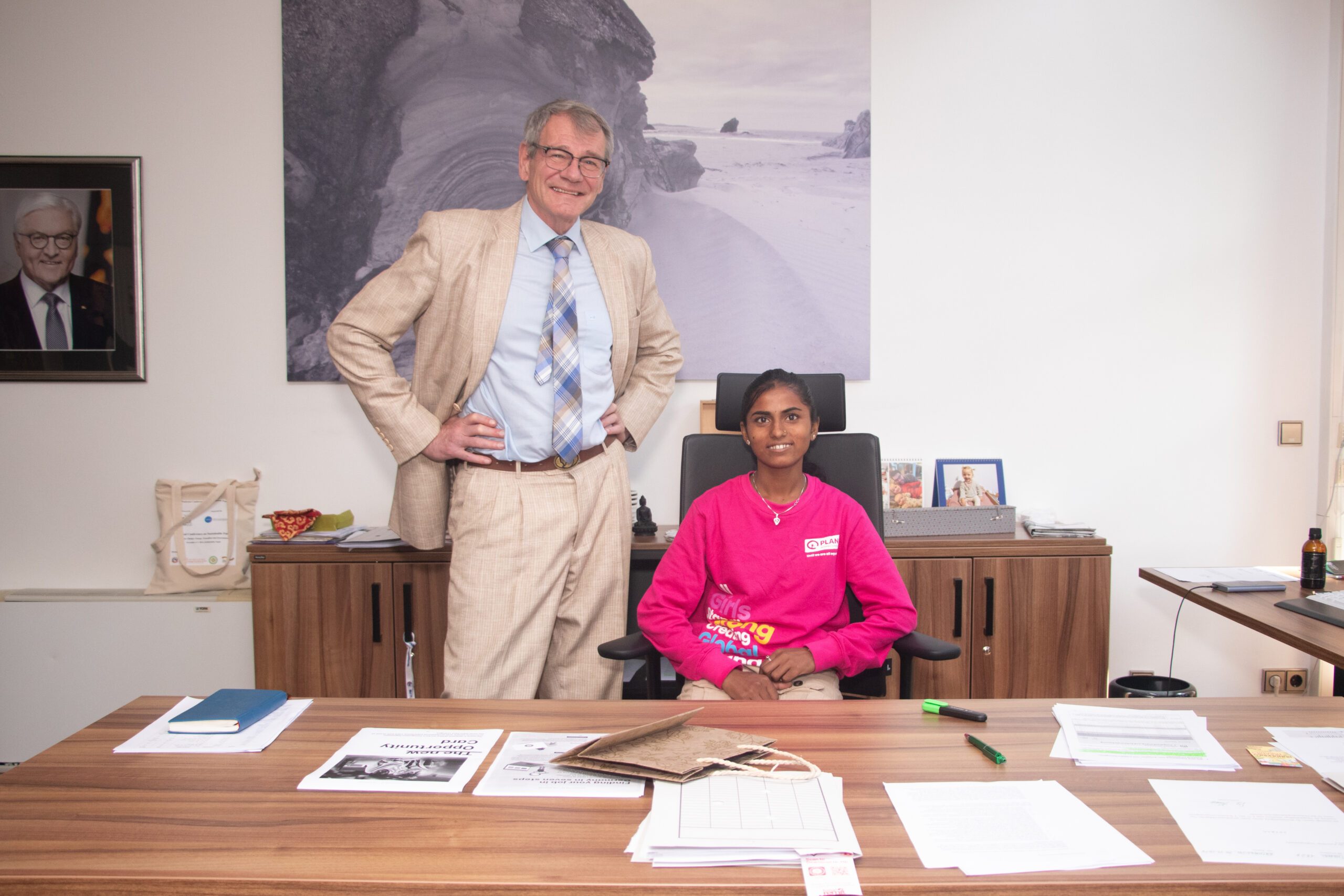 Anita, sitting, wearing pink sweat shirt and H.E. Thomas Prinz German Ambassador standing beside her. 