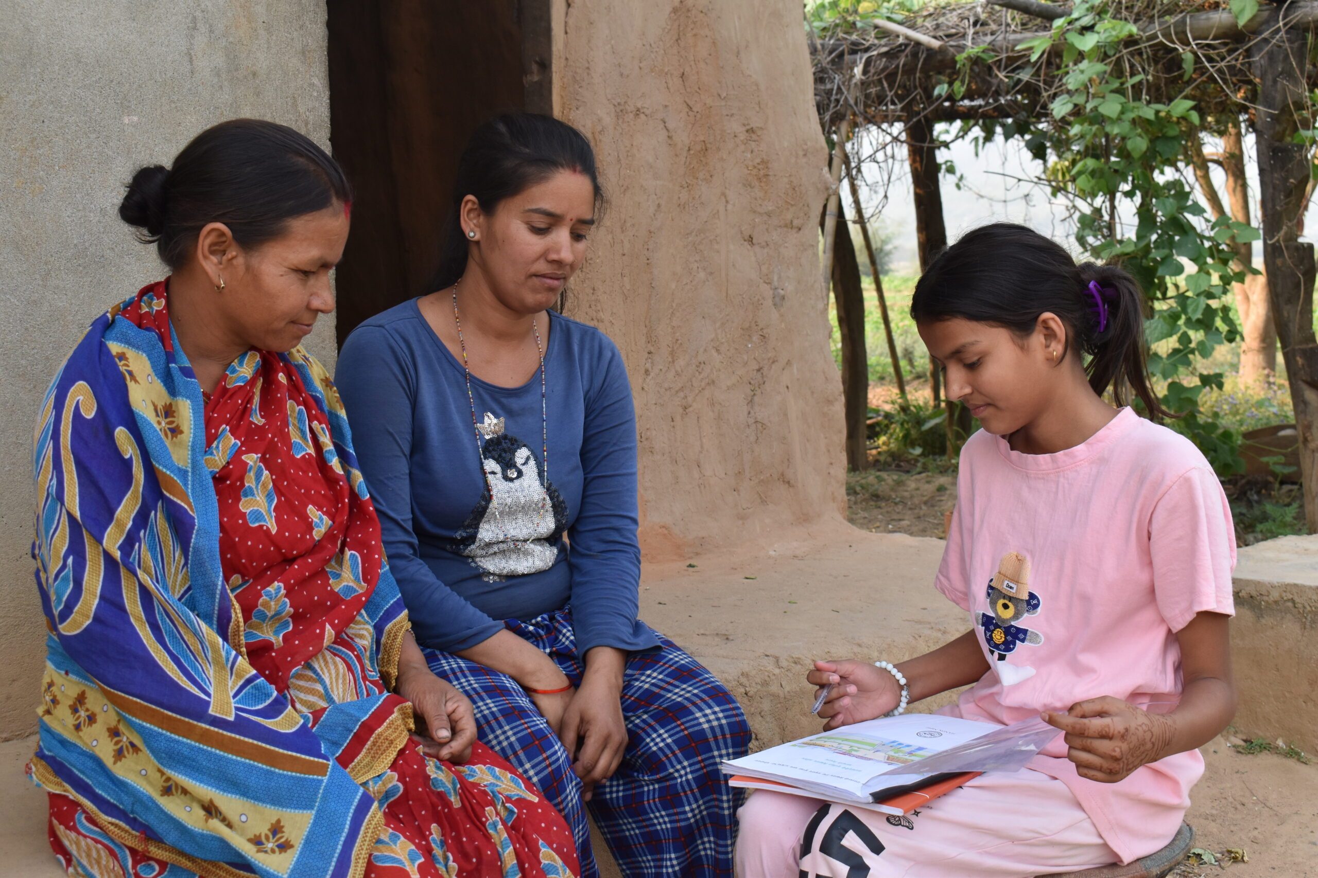 A young girl Sanju with two women sitting outside a house, Sanju holding a book and explaining something to them.