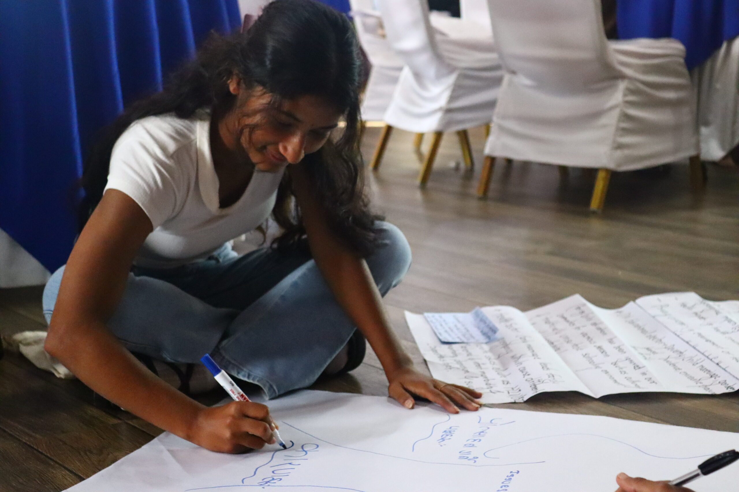 A Young woman with long hair wearing white t-shirt and blue jeans sitting on the floor and writing on the chart paper.
