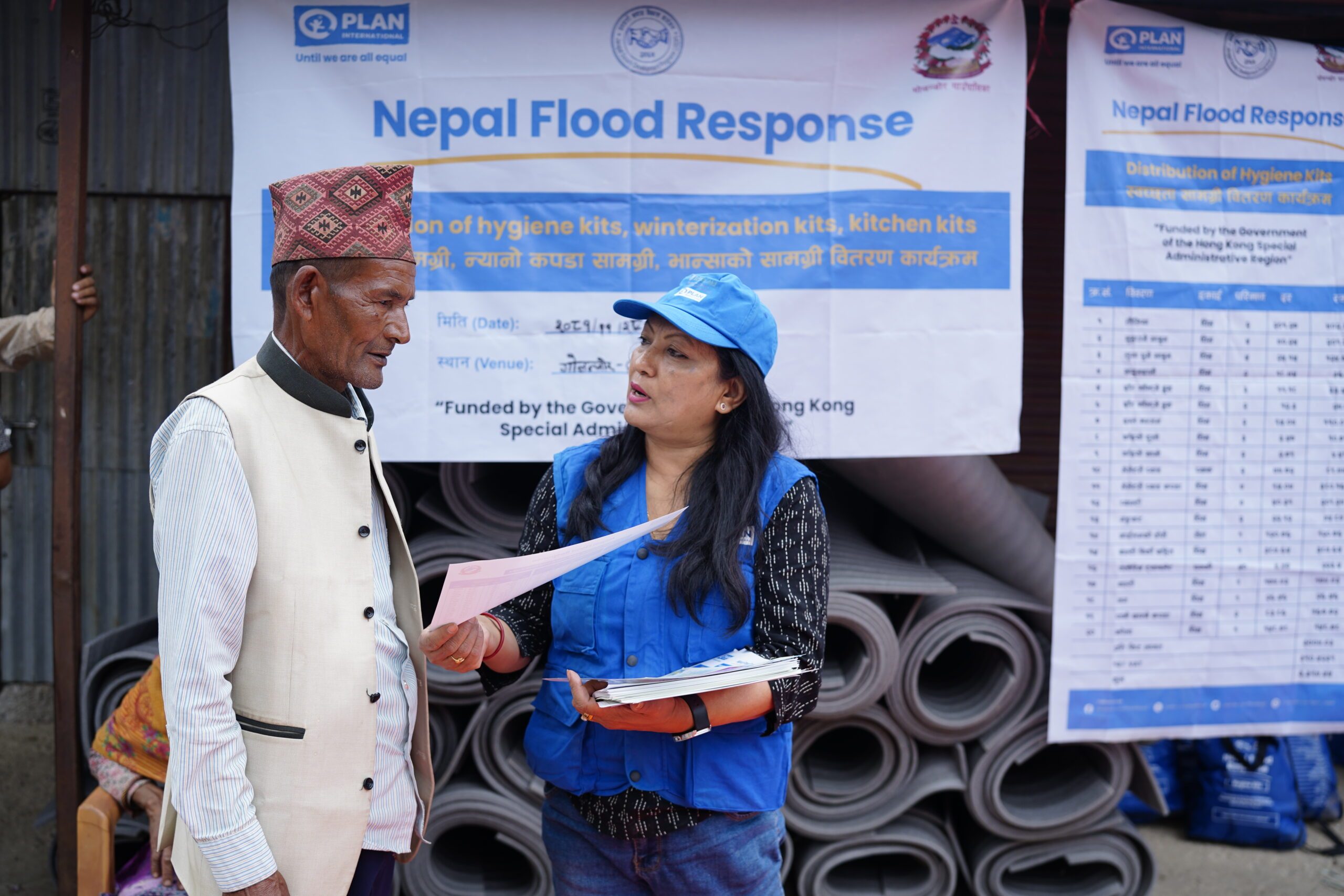 Plan staff wearing blue jacket and cap on right handing over the accountability flyer to a man on left during relief distribution to flood affected in Sindhuli.