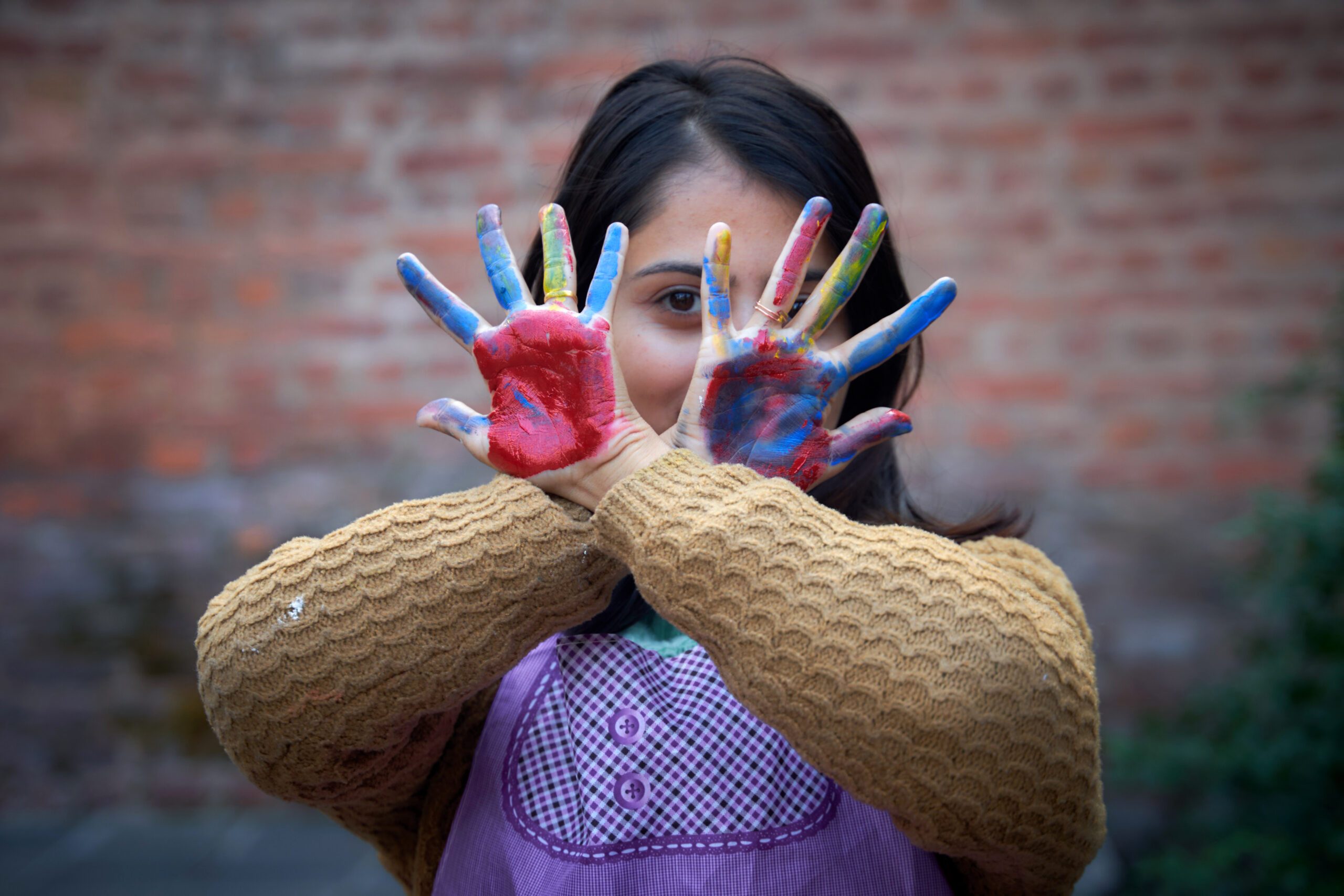 A girl covers her face with her hands painted in bright colours.