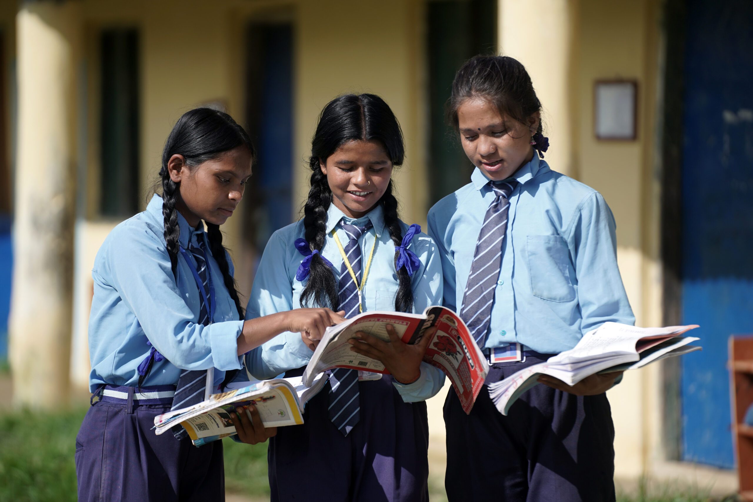 Three girls in school uniform outside their classroom holding books and teaching each other.